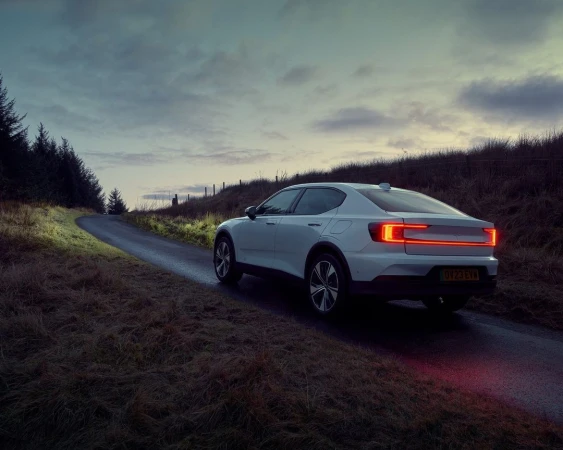 White Polestar 2 electric car on a winding country road at dusk, surrounded by fields and trees, showcasing automotive design and nature.