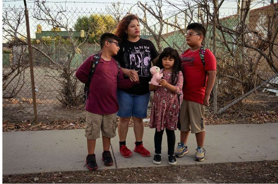 A mother and her three children stand on a sidewalk near a chain-link fence and shrubs. The children wear backpacks, one holds a doll.