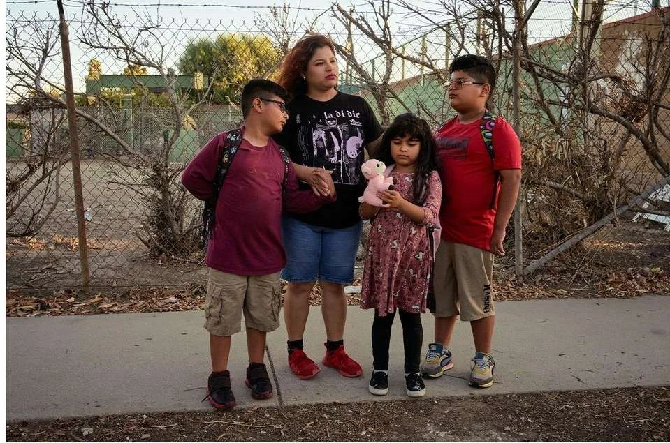A mother and her three children stand on a sidewalk near a chain-link fence and shrubs. The children wear backpacks, one holds a doll.