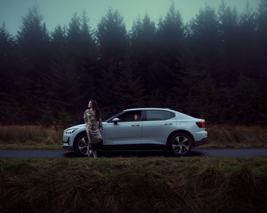 Woman in patterned dress stands next to a white Polestar car in a forest setting, creating a blend of fashion and automotive.