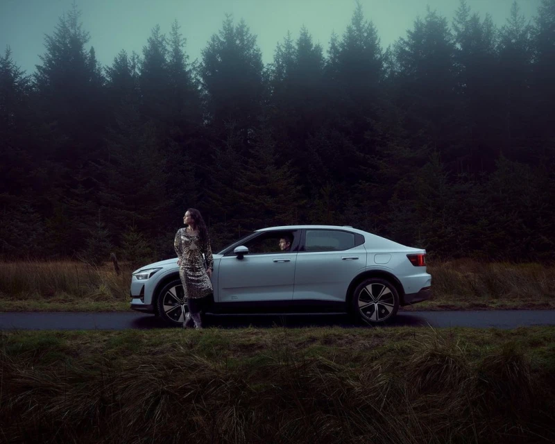 Woman in patterned dress stands next to a white Polestar car in a forest setting, creating a blend of fashion and automotive.