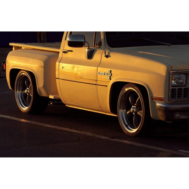 Vintage tan Chevrolet C10 pickup truck in an outdoor setting, featuring custom wheels and lowrider styling in golden light.