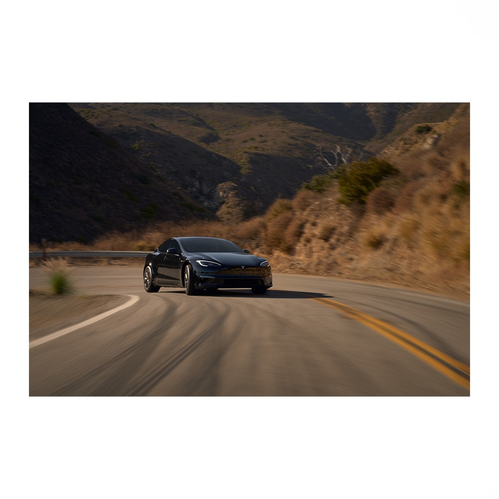A black Tesla Model S driving on a winding mountain road under diffused sunlight, showcasing automotive design and scenic environment.