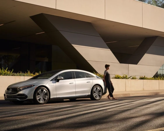 Silver Mercedes-Benz EQS sedan parked on a city street, with an adult leaning against the car. Modern architecture in the background.