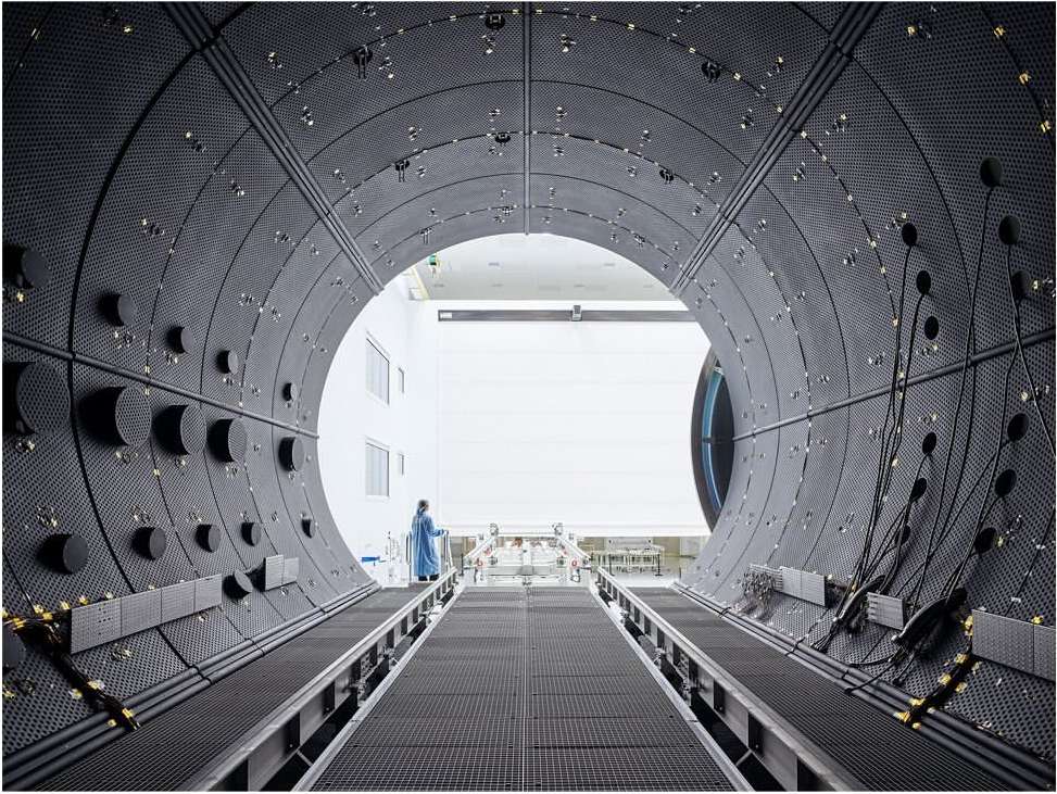 Interior view of a large, dark, tunnel-like industrial space, showing a distant figure in protective wear walking along a pathway.