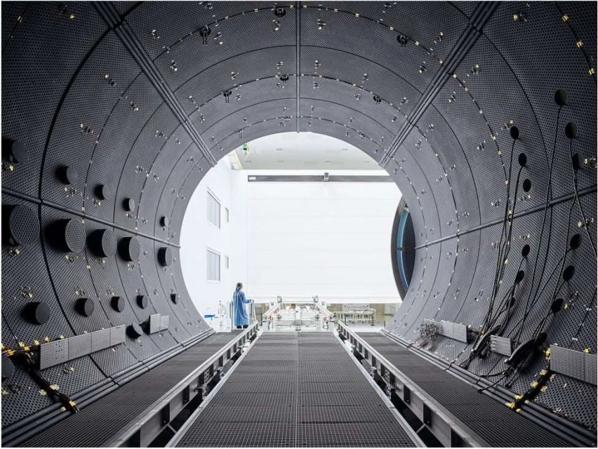 Interior view of a large, dark, tunnel-like industrial space, showing a distant figure in protective wear walking along a pathway.