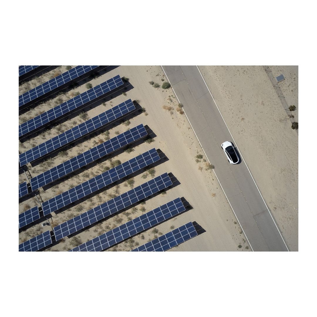 Aerial view of a white electric car driving on a desert road next to a solar farm, showcasing renewable energy and transportation.