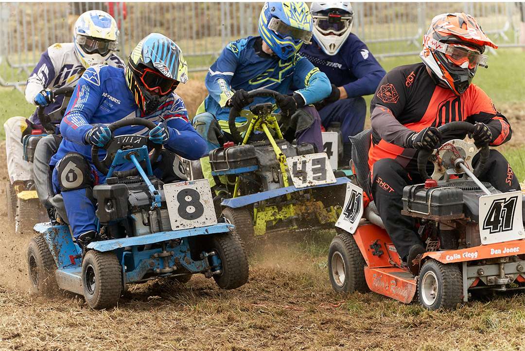 Lawn mower racing event with several racers wearing protective gear competing on a dirt track; the lead racer is in focus.