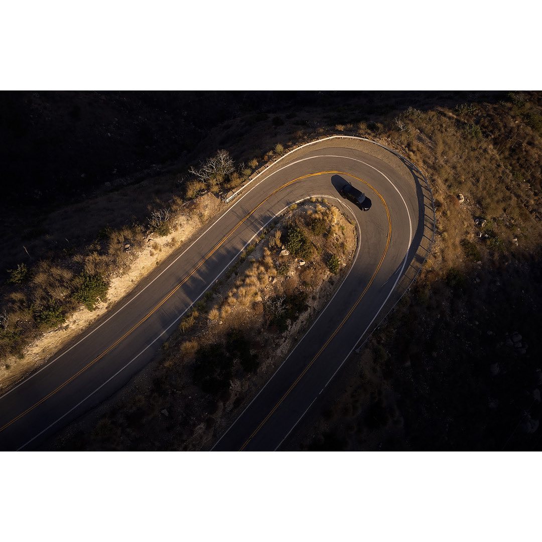 Aerial view of a dark car driving on a winding road through a mountainous landscape during daytime, showcasing travel and automotive.