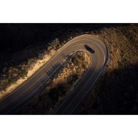 Aerial view of a dark car driving on a winding road through a mountainous landscape during daytime, showcasing travel and automotive.