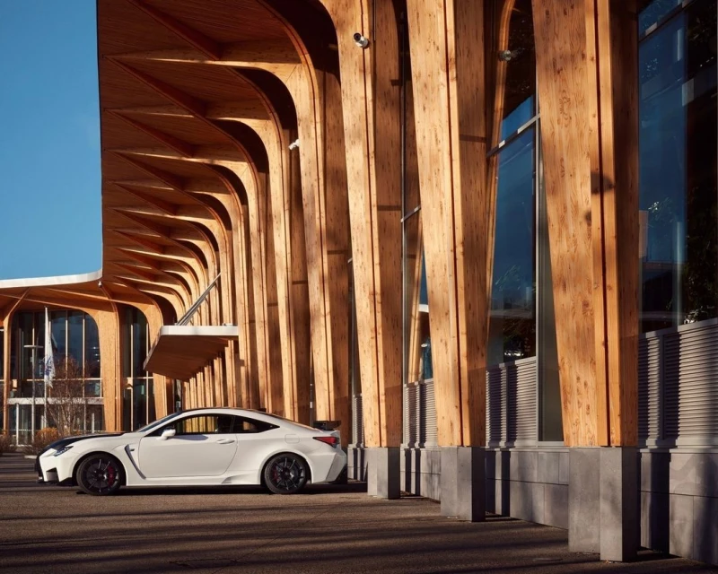 White Lexus RC F parked in front of a modern building featuring light wood architectural beams and large glass windows on a sunny day.