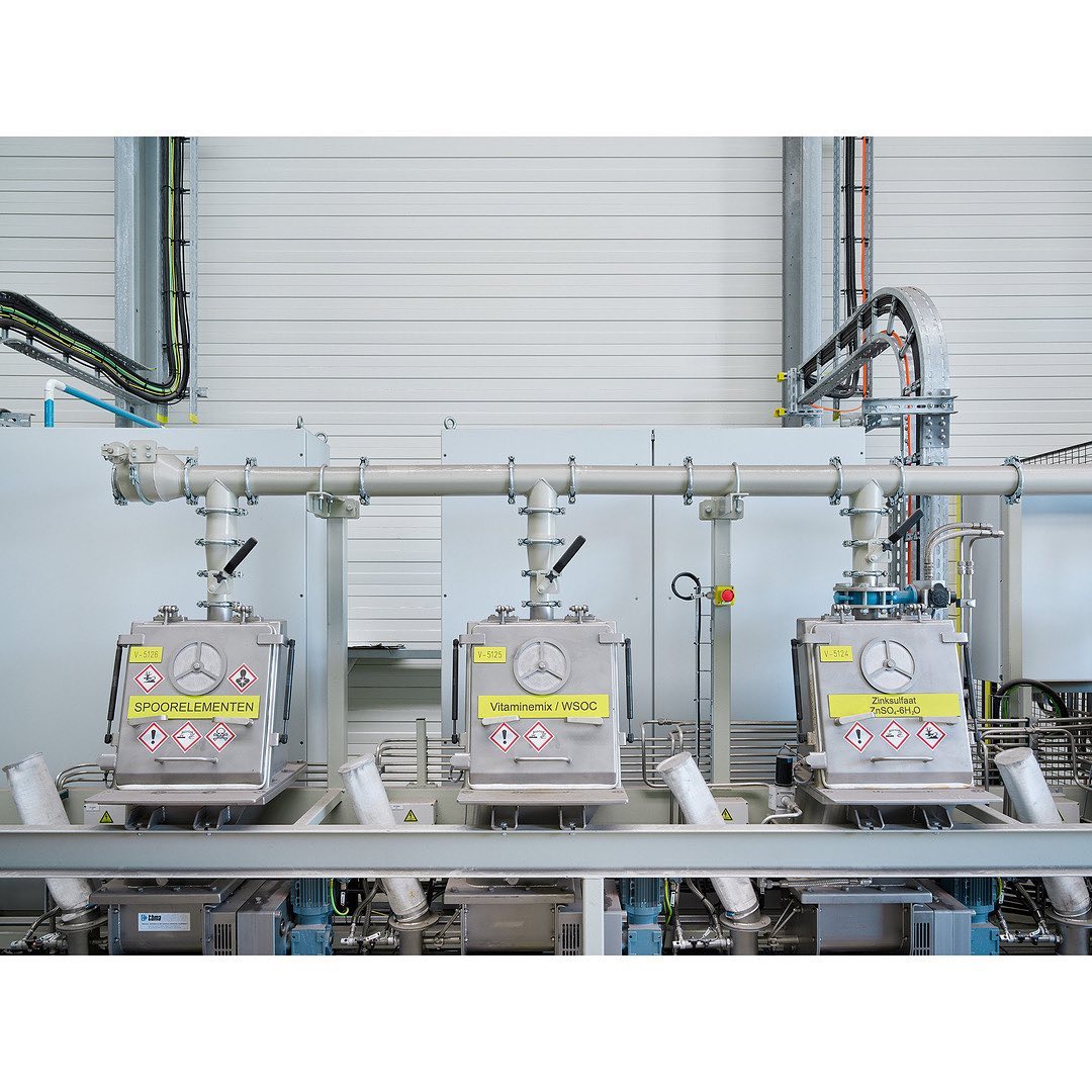 Three industrial stainless steel mixers are connected via pipes against an industrial wall in an indoor factory setting.