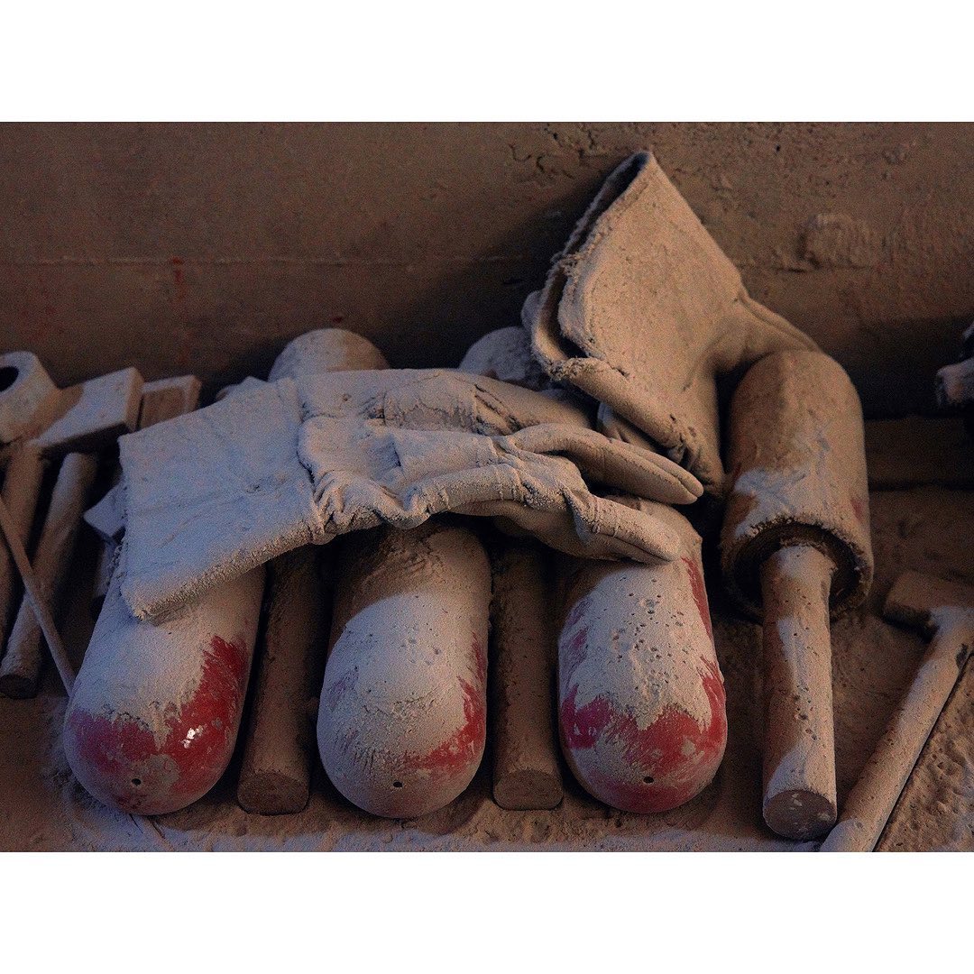Close-up of foundry tools and protective gear covered in sand, including gloves, mallets, and cylindrical objects, with a gray industrial backdrop.