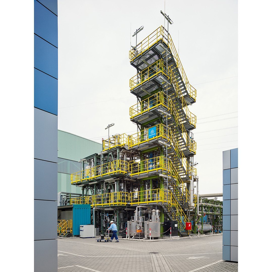 Exterior of an industrial chemical plant complex with scaffolding, machinery, and piping on a sunny day with a worker visible at the base.