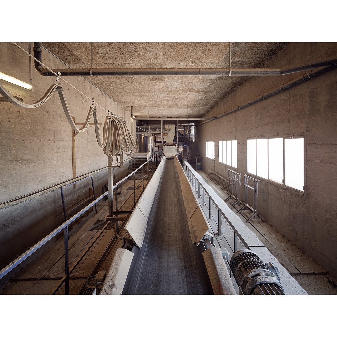 Industrial conveyor belt system in a concrete interior, featuring overhead pipes, rows of windows, and a gray color palette.