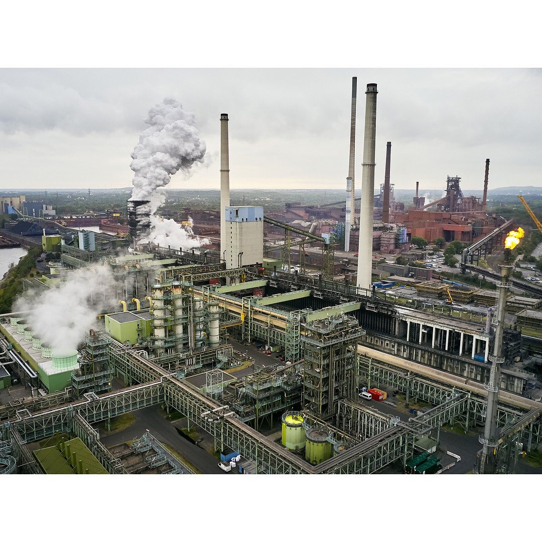 Aerial view of a large industrial complex with multiple chimneys emitting steam under a cloudy sky, showcasing environmental aspects.