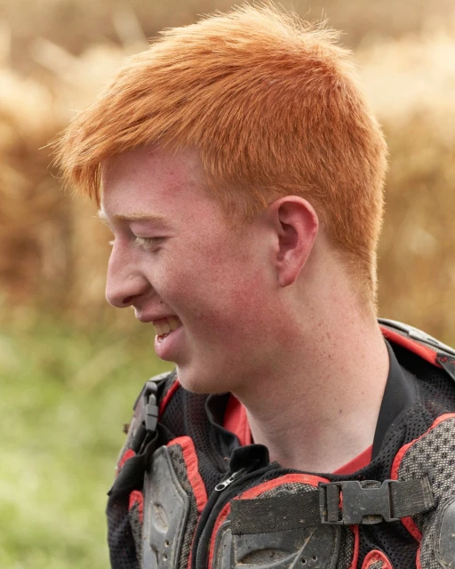 Close-up of a young, smiling redhead wearing protective gear outdoors against a blurred grassy backdrop, showcasing vibrant red hair.