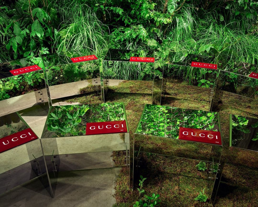 Mirrored planters with Gucci logos are arranged in a lush green garden setting, creating a surreal and reflective outdoor display.