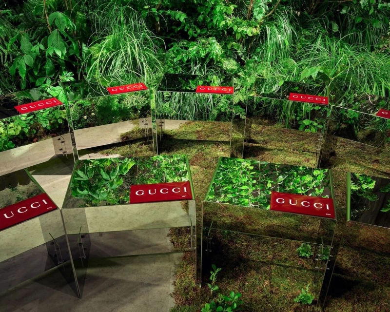 Mirrored planters with Gucci logos are arranged in a lush green garden setting, creating a surreal and reflective outdoor display.