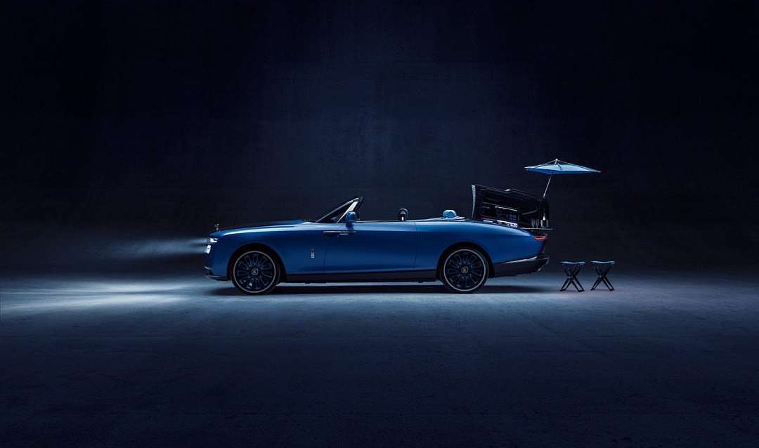 Profile view of a blue Rolls-Royce Boat Tail convertible parked on a dark surface in a studio, with an open trunk and umbrella.