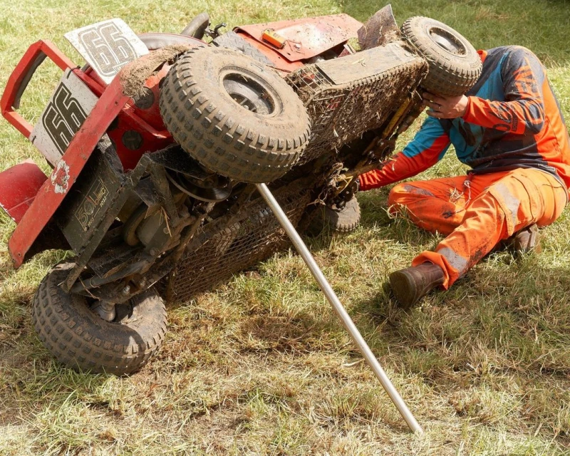 An adult crouches near an overturned red lawnmower on a grassy field, appearing to examine or repair it, on a sunny day.