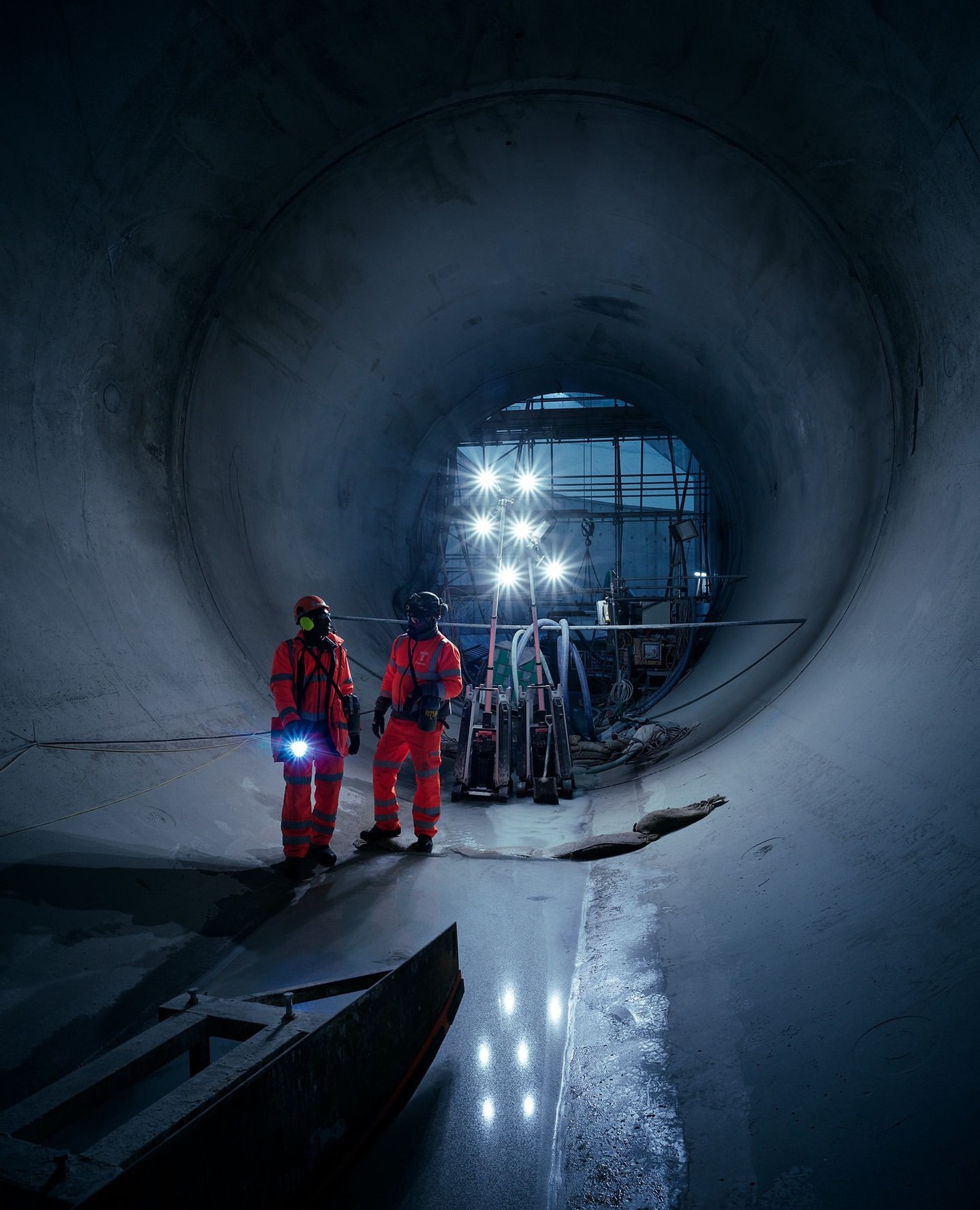 Two construction workers stand inside a large concrete tunnel, illuminated by bright lights and wearing orange safety gear and hard hats.