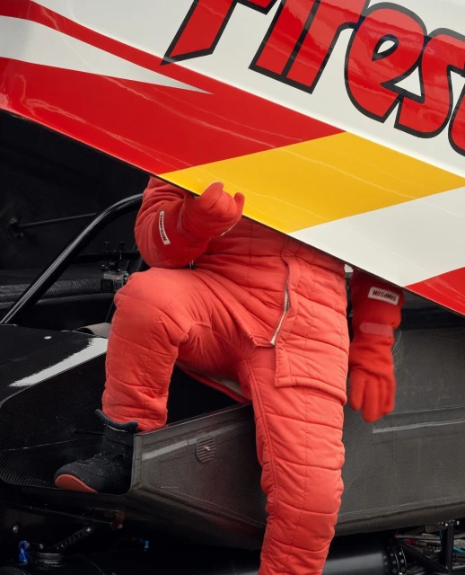 Race car driver in red racing suit climbing into car with Firestone logo on side. Close-up of racer and car, focus on colors.