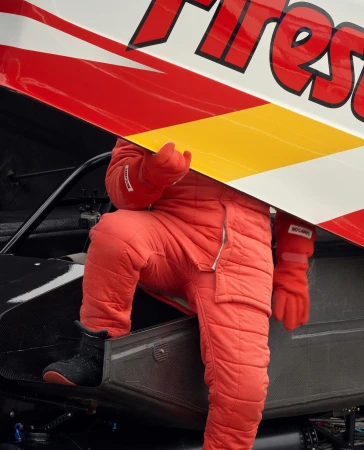 Race car driver in red racing suit climbing into car with Firestone logo on side. Close-up of racer and car, focus on colors.