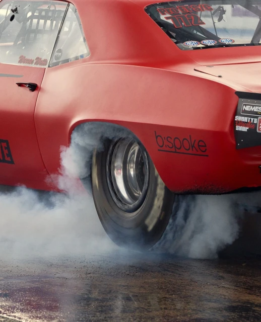 A red race car rear wheel emitting white smoke during a burnout on an asphalt track, car decals and logos visible.