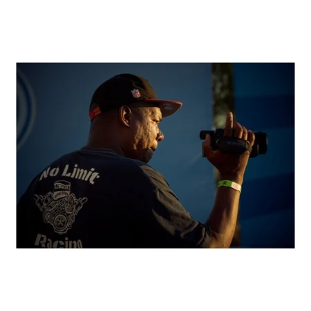 Man films wearing black shirt with "No Limit Racing" engine graphic, cap and wristband. He holds a black camera and looks to the side.