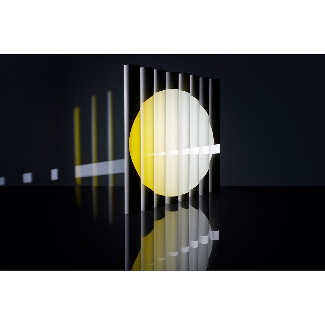 Abstract still life: black vertical bars reflect on surface, backlit by round yellow light. Minimalist design with strong shadow play.