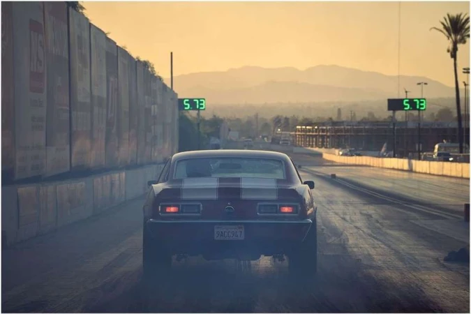 Rear view of a classic muscle car on a drag racing track, with timing displays and mountains in the background during a sunset.