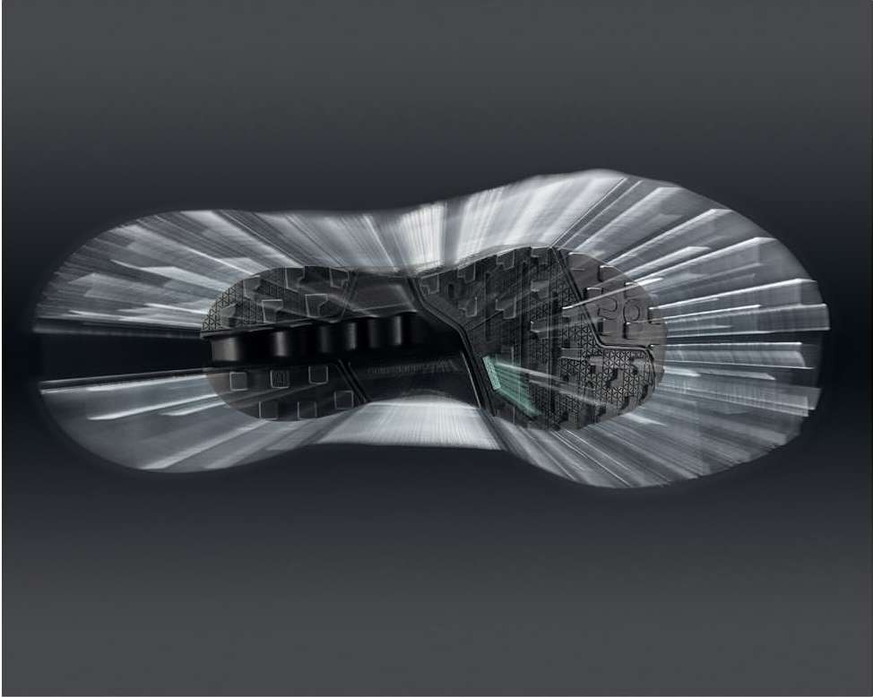 Sole of a black sneaker displayed with a motion effect, showcasing its design and construction against a gradient grey background.