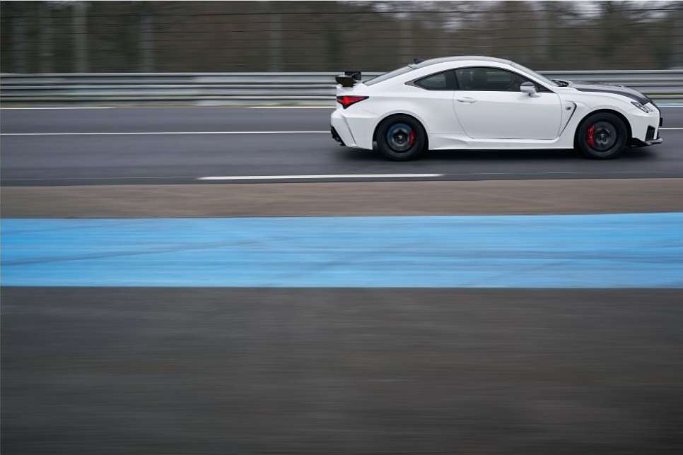 White sports car races along asphalt racetrack with blurred motion; dark grey and blue track lines, daytime, outdoors, automotive racing.