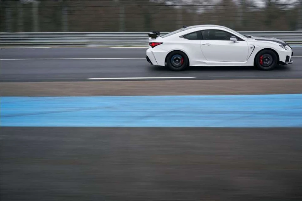 White sports car races along asphalt racetrack with blurred motion; dark grey and blue track lines, daytime, outdoors, automotive racing.