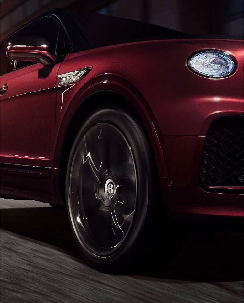 Close-up of a dark red Bentley showcasing its front, wheel, headlight, and body design on a dark road with motion blur.