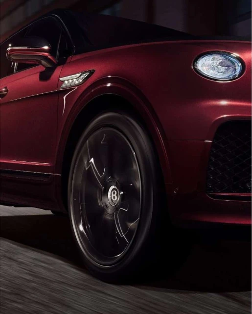 Close-up of a dark red Bentley showcasing its front, wheel, headlight, and body design on a dark road with motion blur.