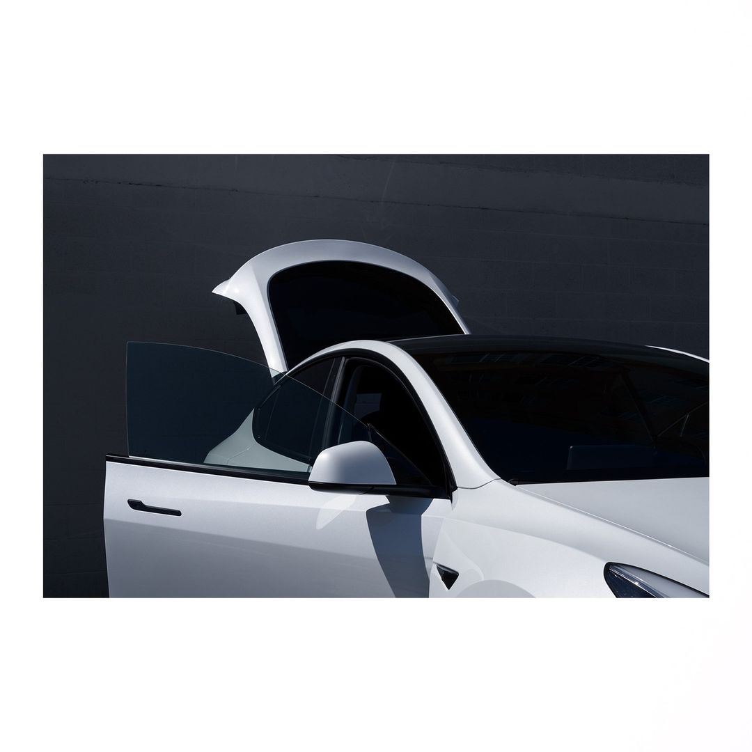 White Tesla Model Y with open rear door and rear hatch, showcased against a dark gray background in a modern automotive setting.