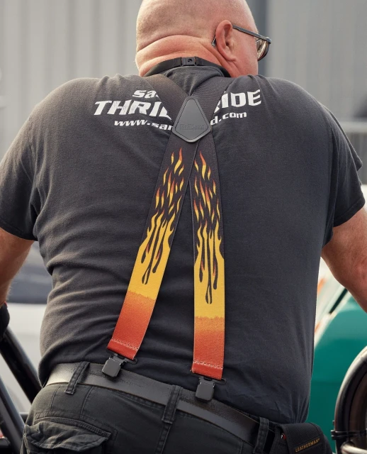 Man wearing suspenders with flame design, black t-shirt, and jeans stands outdoors. The back of the person is visible.
