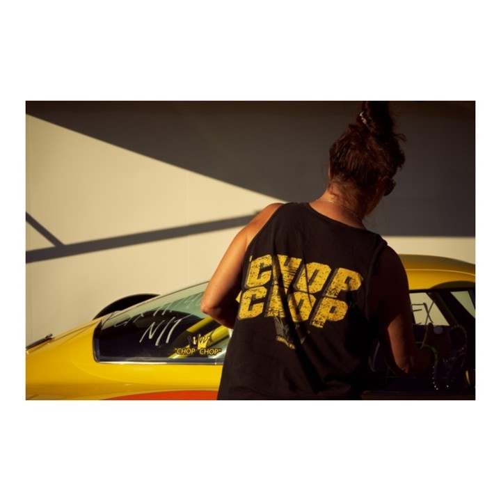 A person stands next to a yellow drag racing car, wearing a black tank top with graphic text. Exterior daylight scene with shadows.