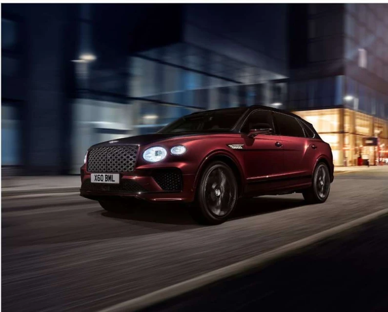 Burgundy Bentley SUV on the road at night, blurred motion, city lights visible in background, showcasing automotive luxury and design.
