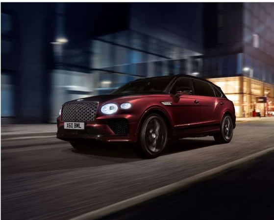 Burgundy Bentley SUV on the road at night, blurred motion, city lights visible in background, showcasing automotive luxury and design.