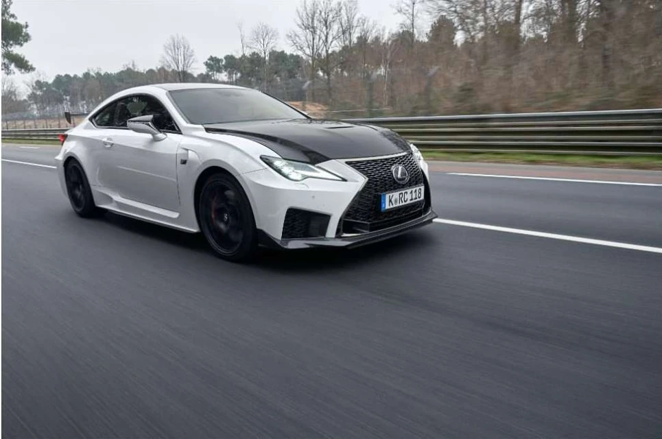 White Lexus RC F driving on a racetrack with blurred motion effect, showcasing its design and speed in an outdoor setting.