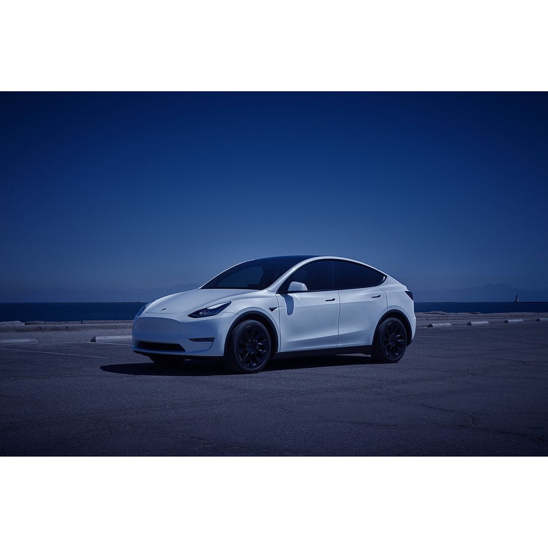 Tesla Model Y parked near the coast under a blue sky, showcasing its sleek design and the vehicle's presence in a scenic outdoor setting.