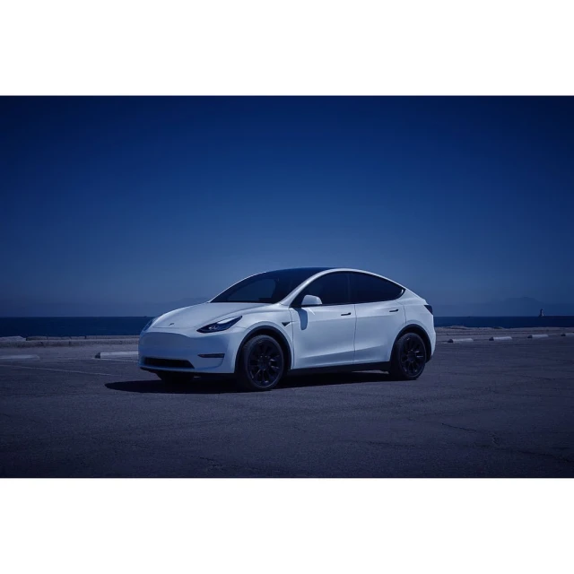 Tesla Model Y parked near the coast under a blue sky, showcasing its sleek design and the vehicle's presence in a scenic outdoor setting.