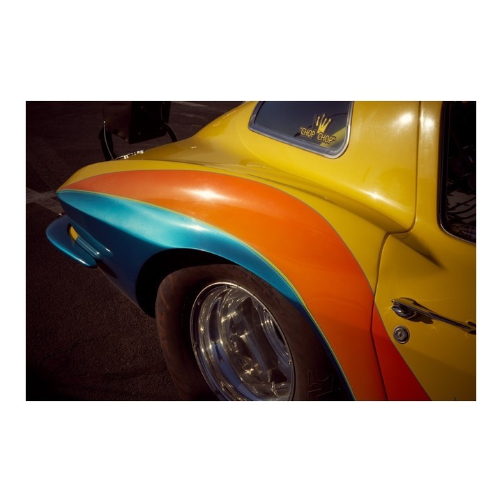 A yellow vintage car with blue, orange, and yellow paint stripes over the rear fender and a chrome wheel, shot in diffused sunlight.