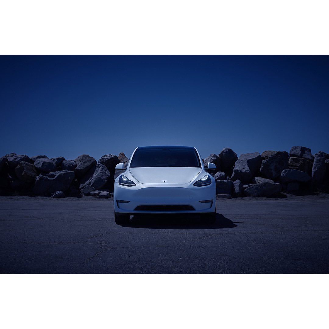Front view of a white Tesla car parked in an outdoor lot with a rock wall and blue sky in the background, car facing camera.