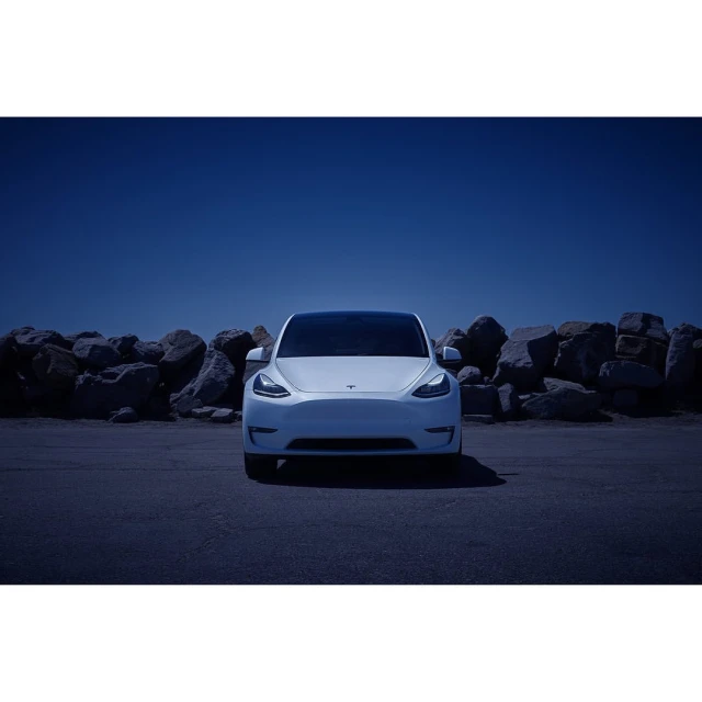 Front view of a white Tesla car parked in an outdoor lot with a rock wall and blue sky in the background, car facing camera.