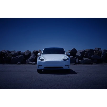 Front view of a white Tesla car parked in an outdoor lot with a rock wall and blue sky in the background, car facing camera.