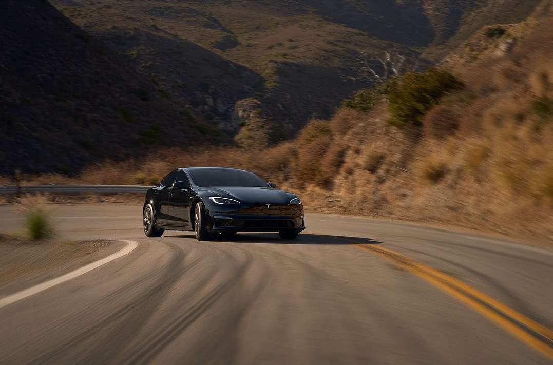 Black electric car drives on asphalt road lined with golden grass, backed by arid hills under a bright sunny sky, in an automotive setting.
